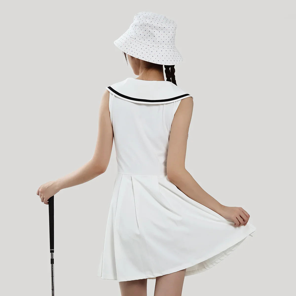 Woman wearing a white dress with black trim and a matching hat, holding a cane on a plain background
