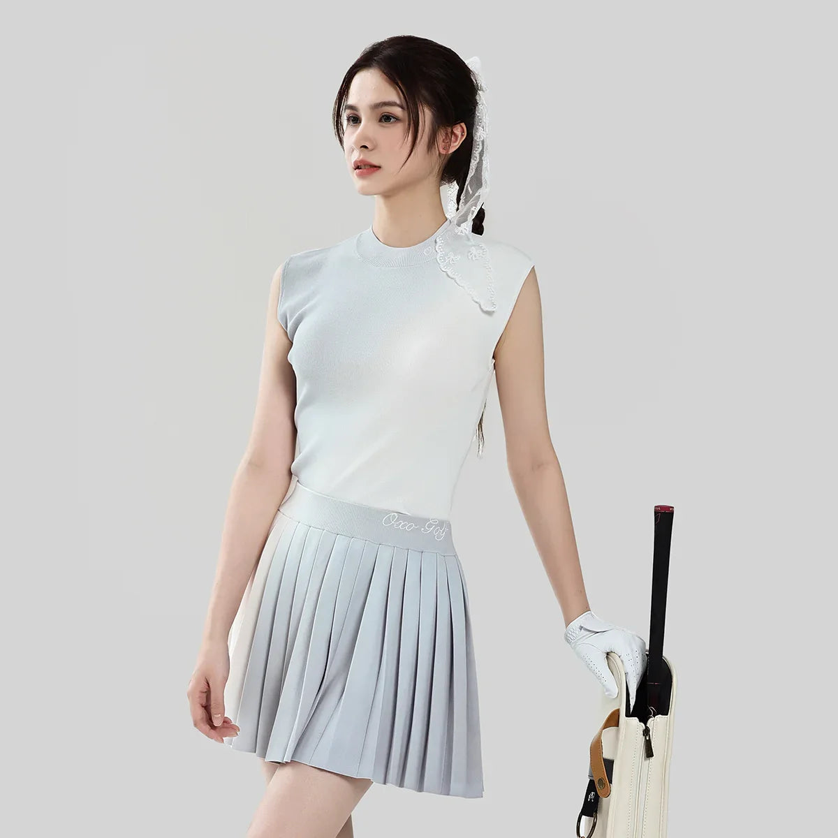 Woman in light gray sleeveless top and pleated skirt holding a golf club on a plain background