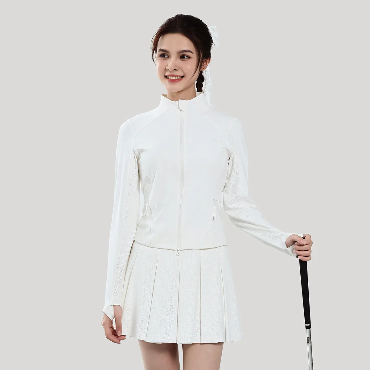 Woman wearing a white outfit with a pleated skirt, holding a golf club on a plain background