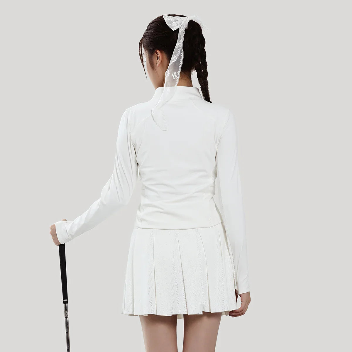 Person wearing a white outfit with a pleated skirt and long-sleeve top, holding a tennis racket on a plain background.