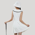 Woman wearing a white dress with black trim and a matching hat, holding a cane on a plain background